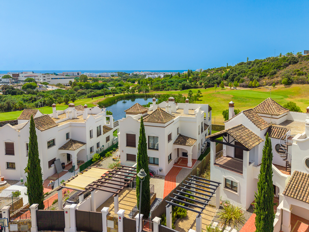 High-angle view of white Mediterranean-style villas with tiled roofs, surrounded by lush greenery, a pond, and the ocean.