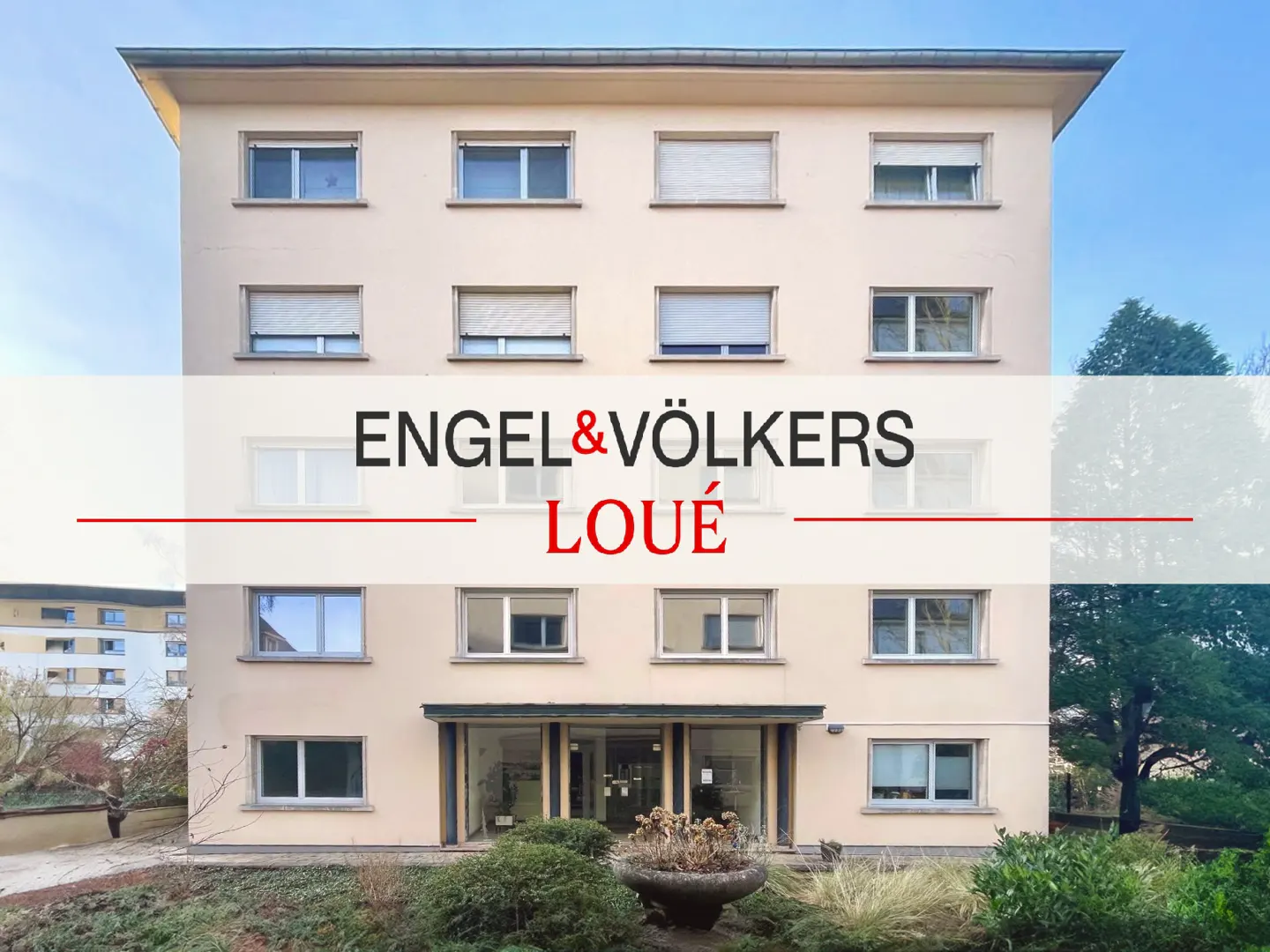 Three-story apartment building with beige facade and many windows. Engel & Völkers logo overlayed. Front garden with greenery.