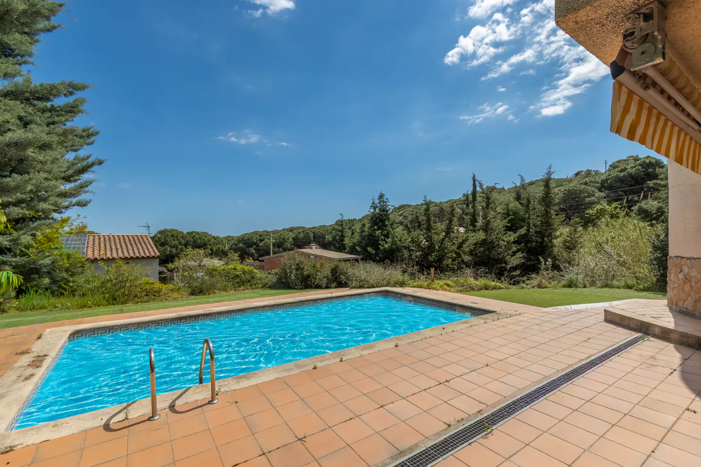Outdoor pool with blue water, surrounded by terracotta tiles and green grass. Trees and a blue sky with clouds in the background.