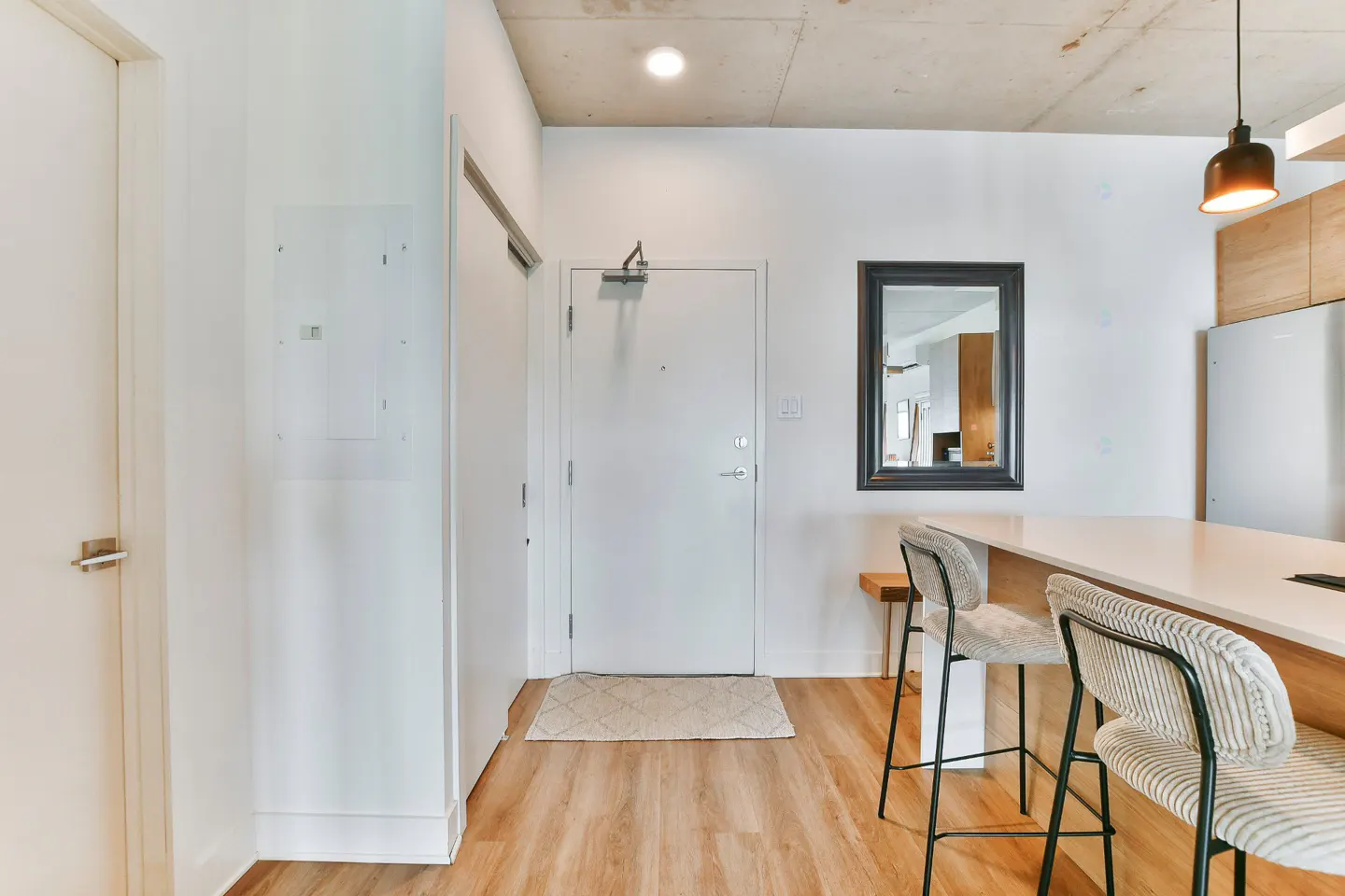 Modern apartment entryway with white walls, wood floors, a white door, a mirror, and a kitchen island with two bar stools.