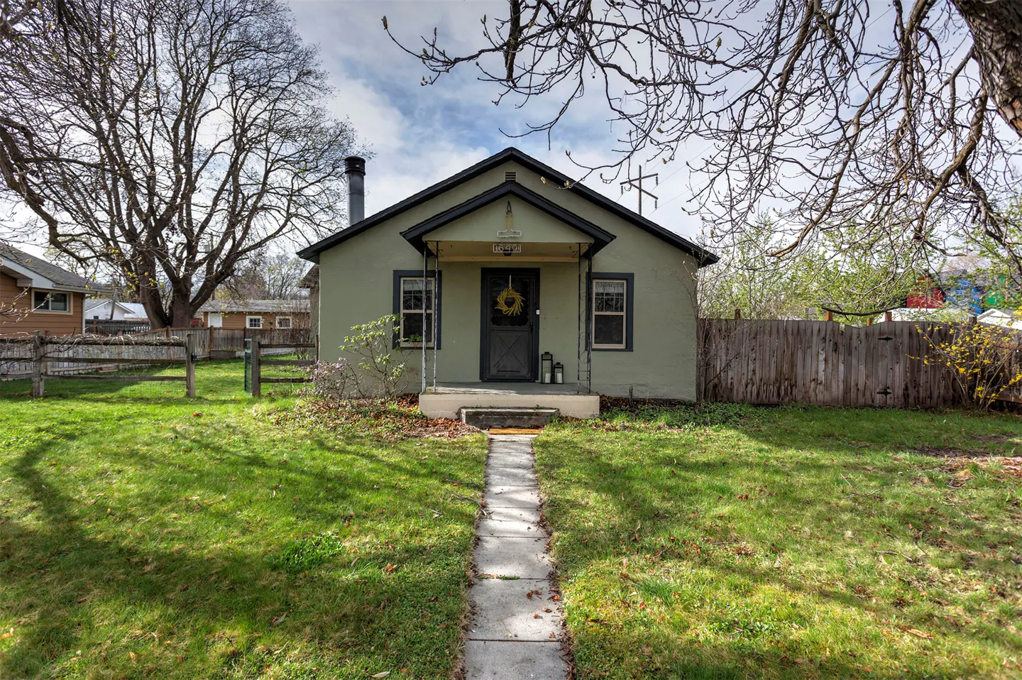 A quaint, light green bungalow with a black door and wreath, framed by a green lawn and trees.