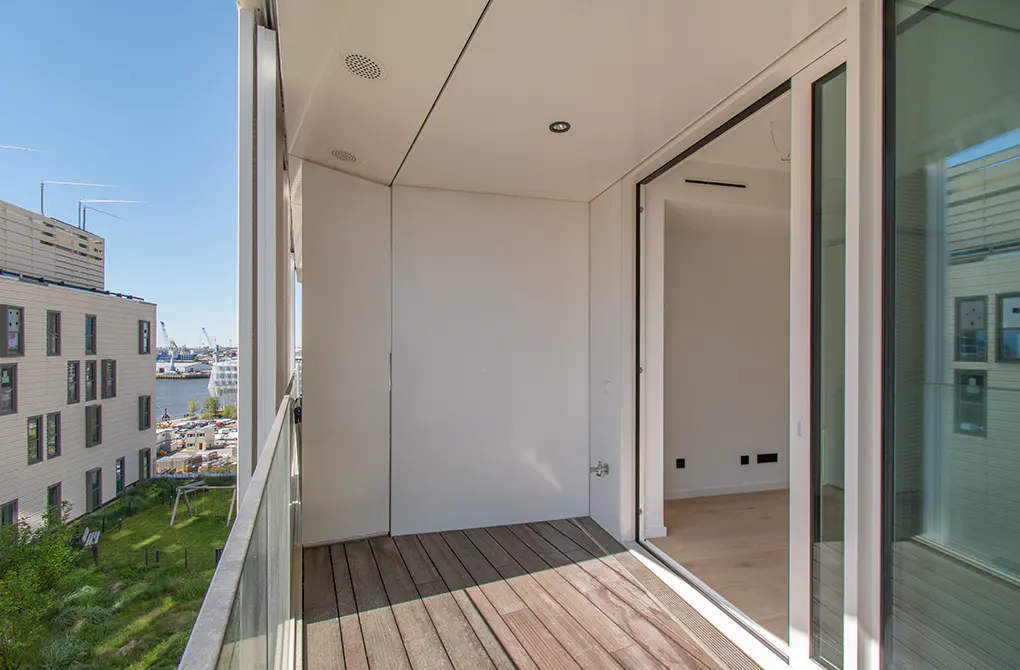 Modern balcony with wooden flooring and glass railing, overlooking a harbor view with nearby buildings and greenery.