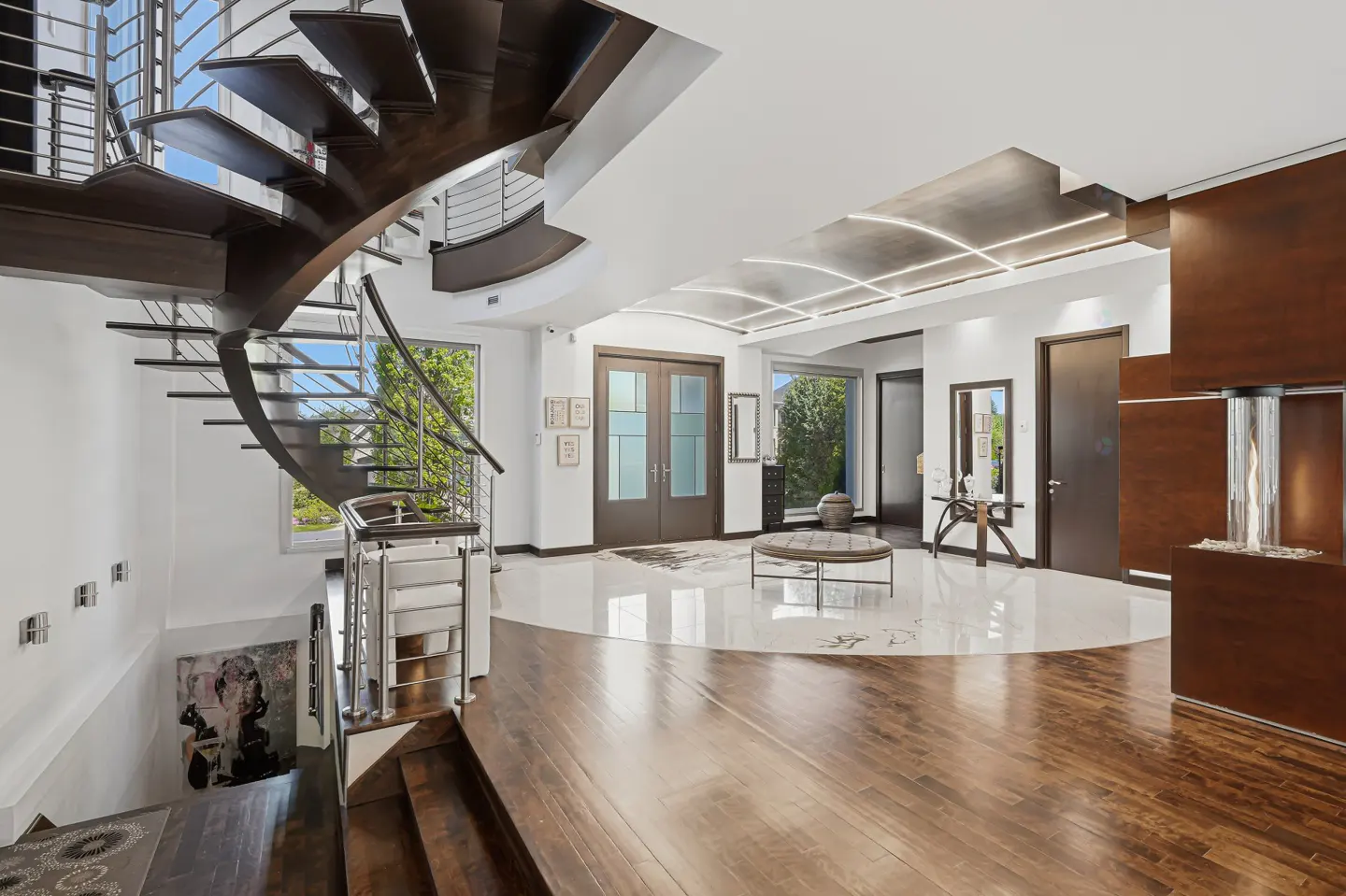 Bright foyer with a dark wood spiral staircase, stainless steel railings, and a white marble floor. Double doors lead to the outside.