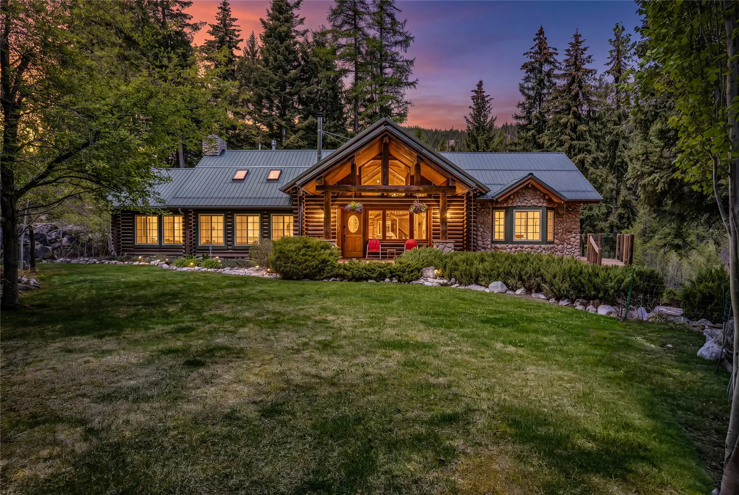 Log cabin home with green roof, surrounded by trees and green lawn at dusk. Lights are on inside.