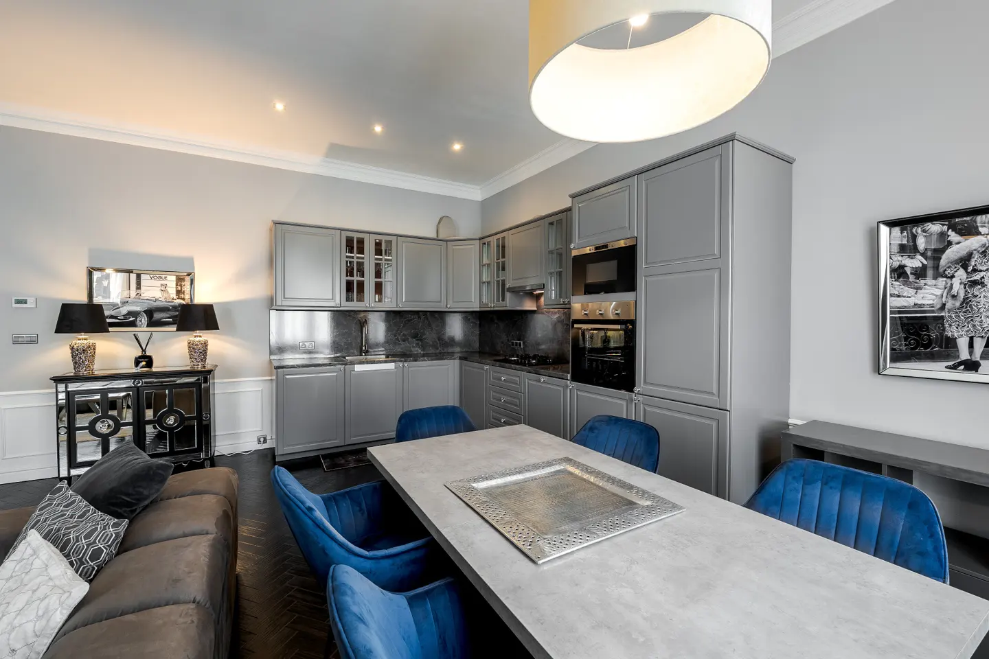 A modern kitchen and dining area with gray cabinets, a concrete table with blue velvet chairs, and a gray sofa.