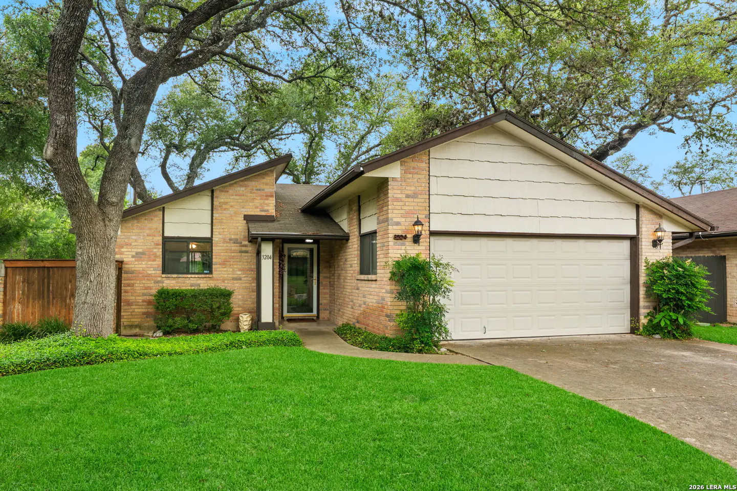 A single-story brick house with a white garage door and green lawn under a blue sky with trees.