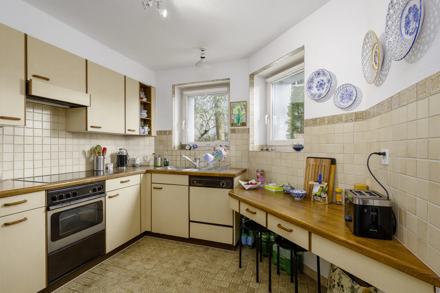 A bright kitchen with beige cabinets, a black oven, and a wooden countertop. Decorative plates adorn the wall. Two windows offer natural light.