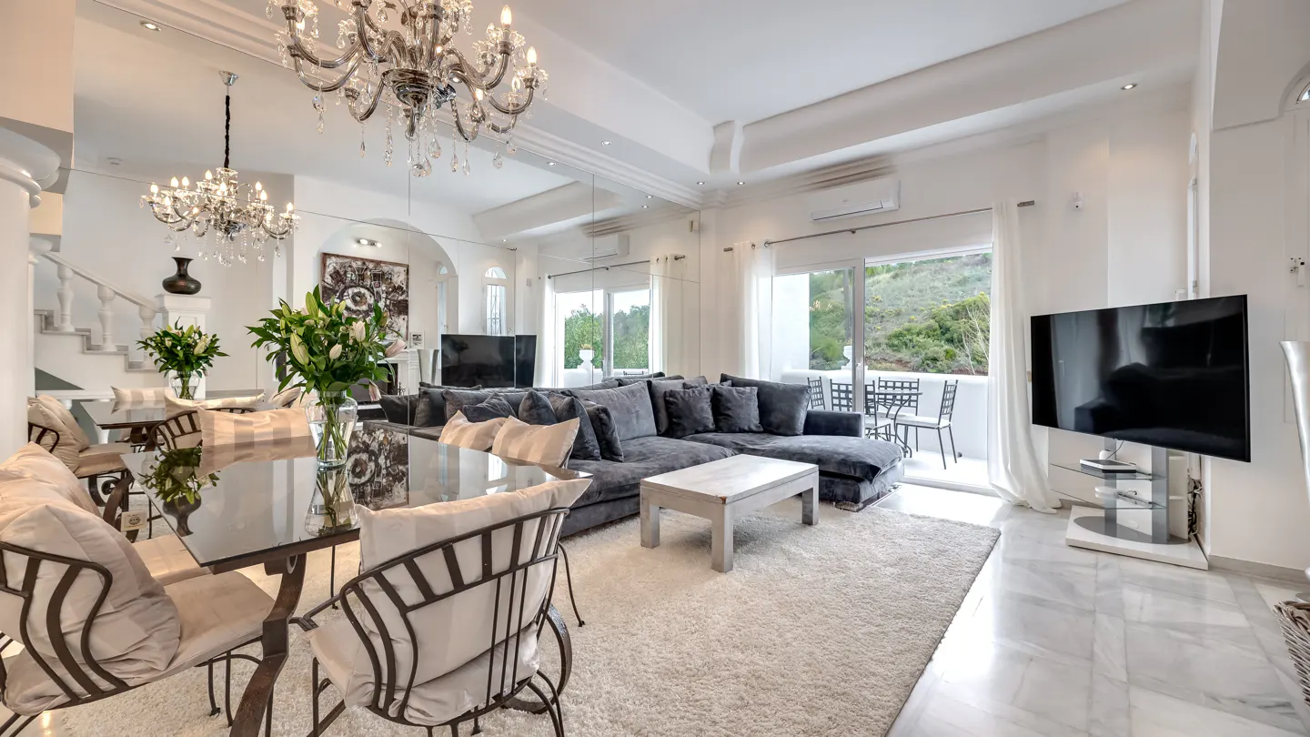 Bright living room with a gray sectional sofa, glass table, and crystal chandelier. Balcony with chairs visible through sliding glass doors.
