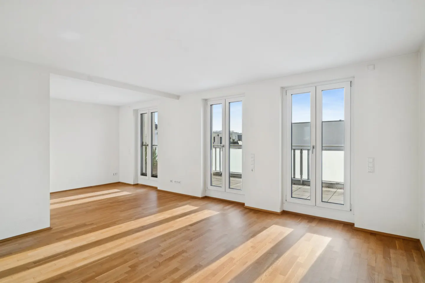 Bright, empty room with hardwood floors, white walls, and three white-framed glass doors leading to a balcony.
