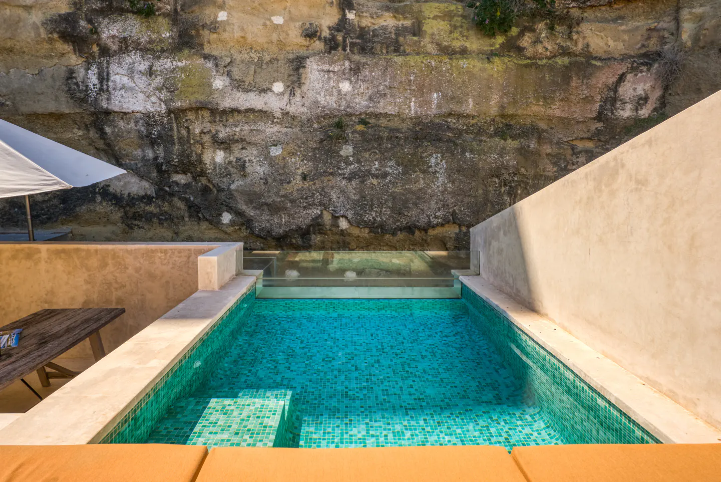 A turquoise tiled pool is nestled against a stone wall, with a table, umbrella, and lounge chairs nearby.