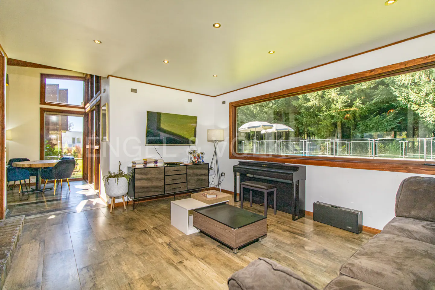 Living room with wood floors, a TV, a piano, and a large window overlooking a pool and trees.