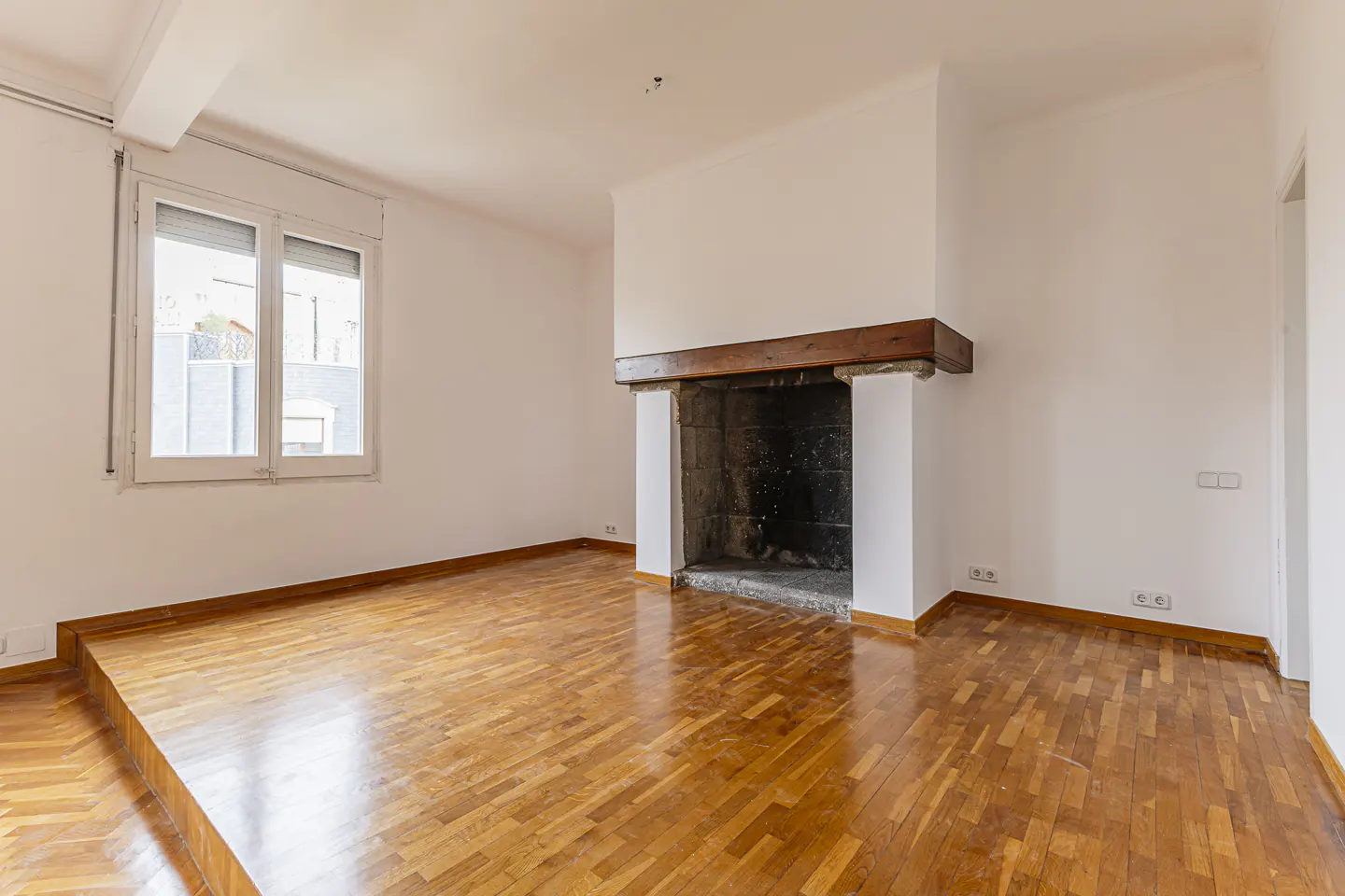 Empty room with polished wood floors, white walls, a large window, and a rustic stone fireplace with a wooden mantel.