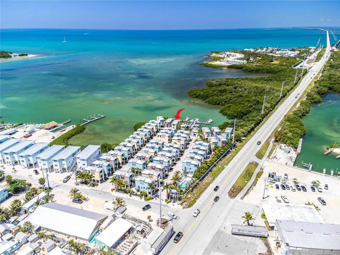 Aerial view of colorful waterfront homes with docks on turquoise water, next to a highway and lush greenery under a clear blue sky.