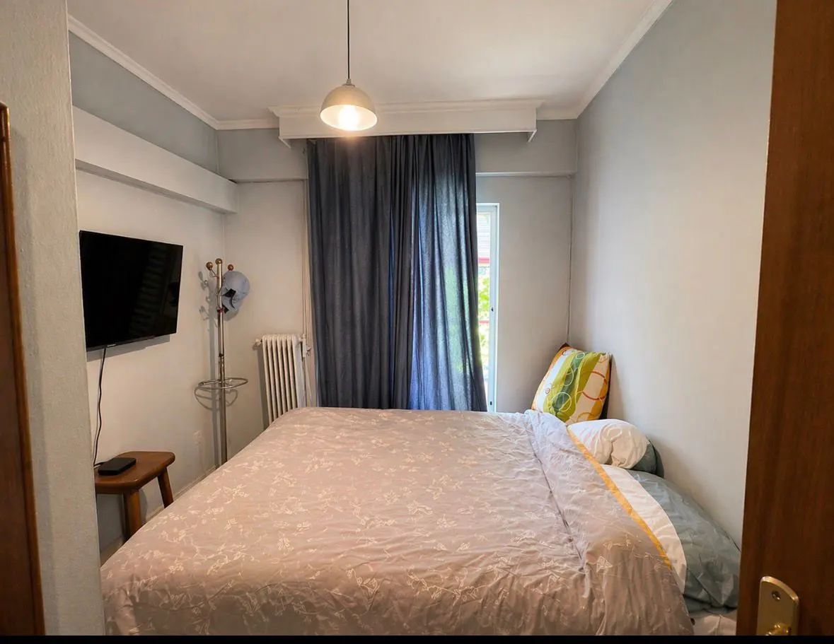 Bedroom with a double bed, TV, coat rack, radiator, and blue curtains. The walls are painted light gray.