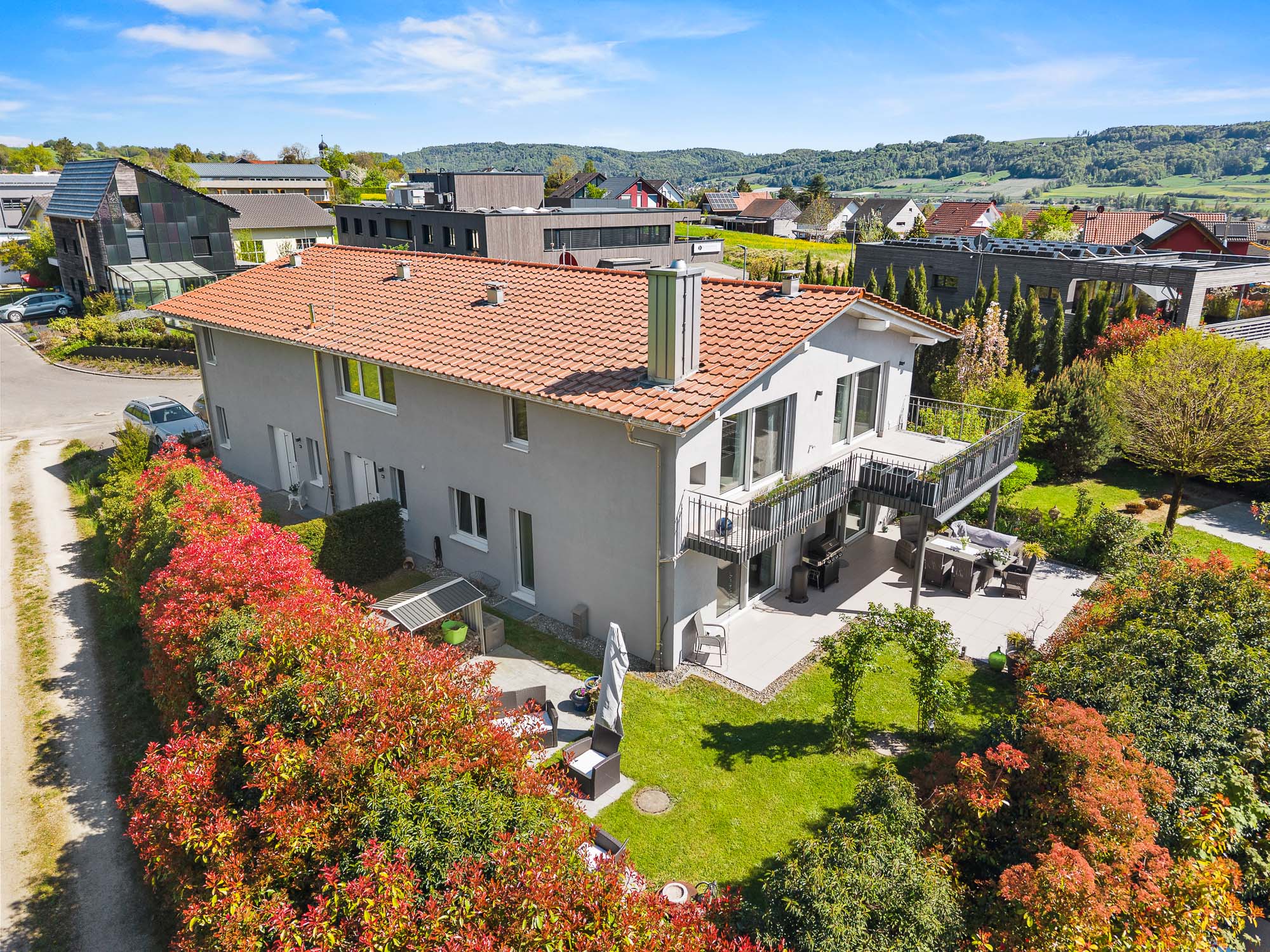 Aerial view of a two-story gray house with a red tile roof, balcony, patio, and lush garden in a suburban neighborhood.