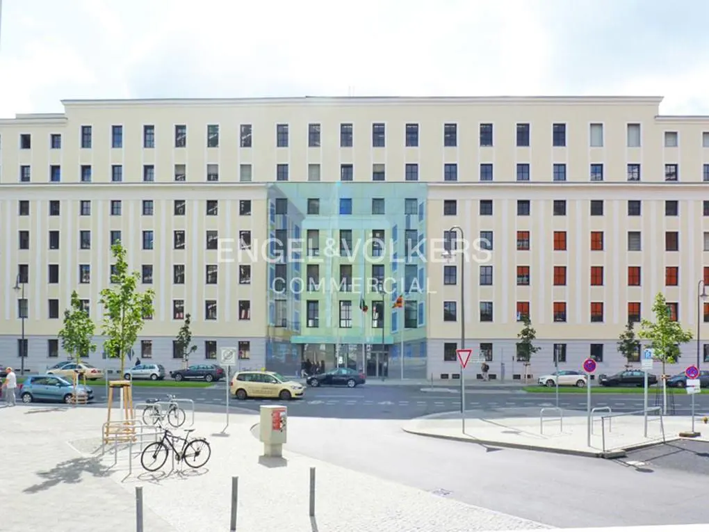 Exterior of a beige building with "Engel & Völkers Commercial" on a glass facade. Cars and bikes are parked nearby.
