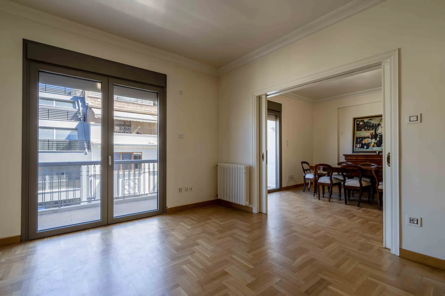 Bright, empty room with herringbone wood floors, a balcony with gray doors, and a dining room visible through a doorway.
