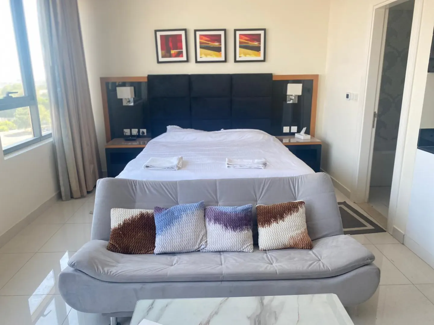 A modern hotel room features a queen bed with a black headboard, a gray sofa with colorful pillows, and a marble coffee table.
