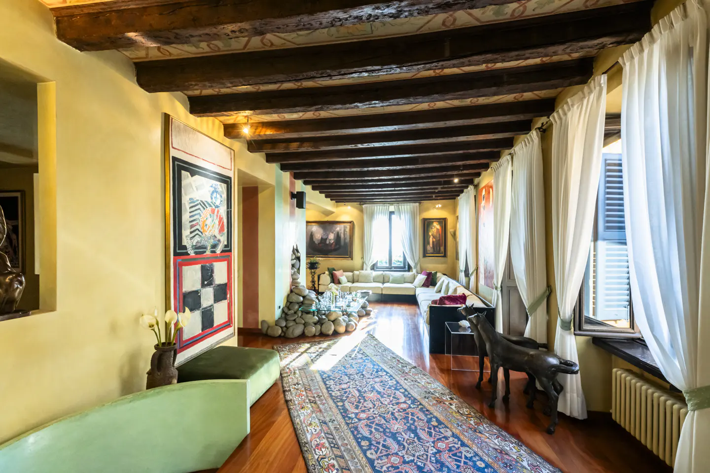 A living room with yellow walls, wood floors, and a beamed ceiling. A blue rug runs through the center. Art and sculptures decorate the space.