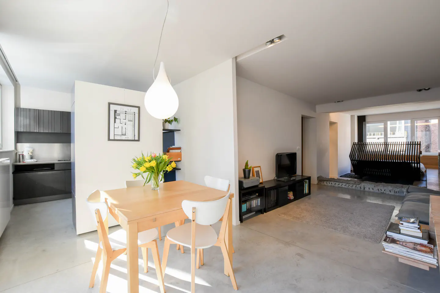 Bright, open-concept apartment with a dining area featuring a wooden table, white chairs, and a vase of yellow tulips. A kitchen and living room are visible.