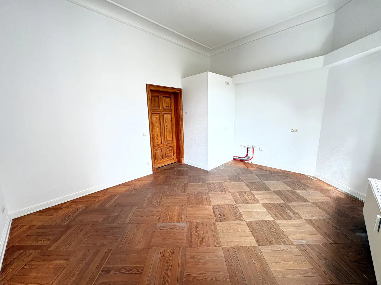 Empty room with white walls, a brown parquet floor, and a wooden door on the left. Red pipes are visible on the right wall.