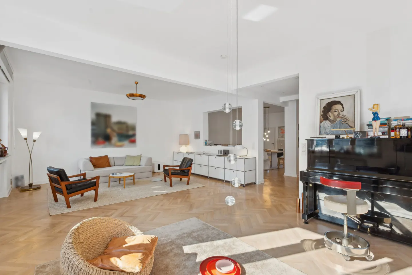 Bright, open living room with herringbone floors, white walls, and modern furniture. A black piano sits near a portrait.