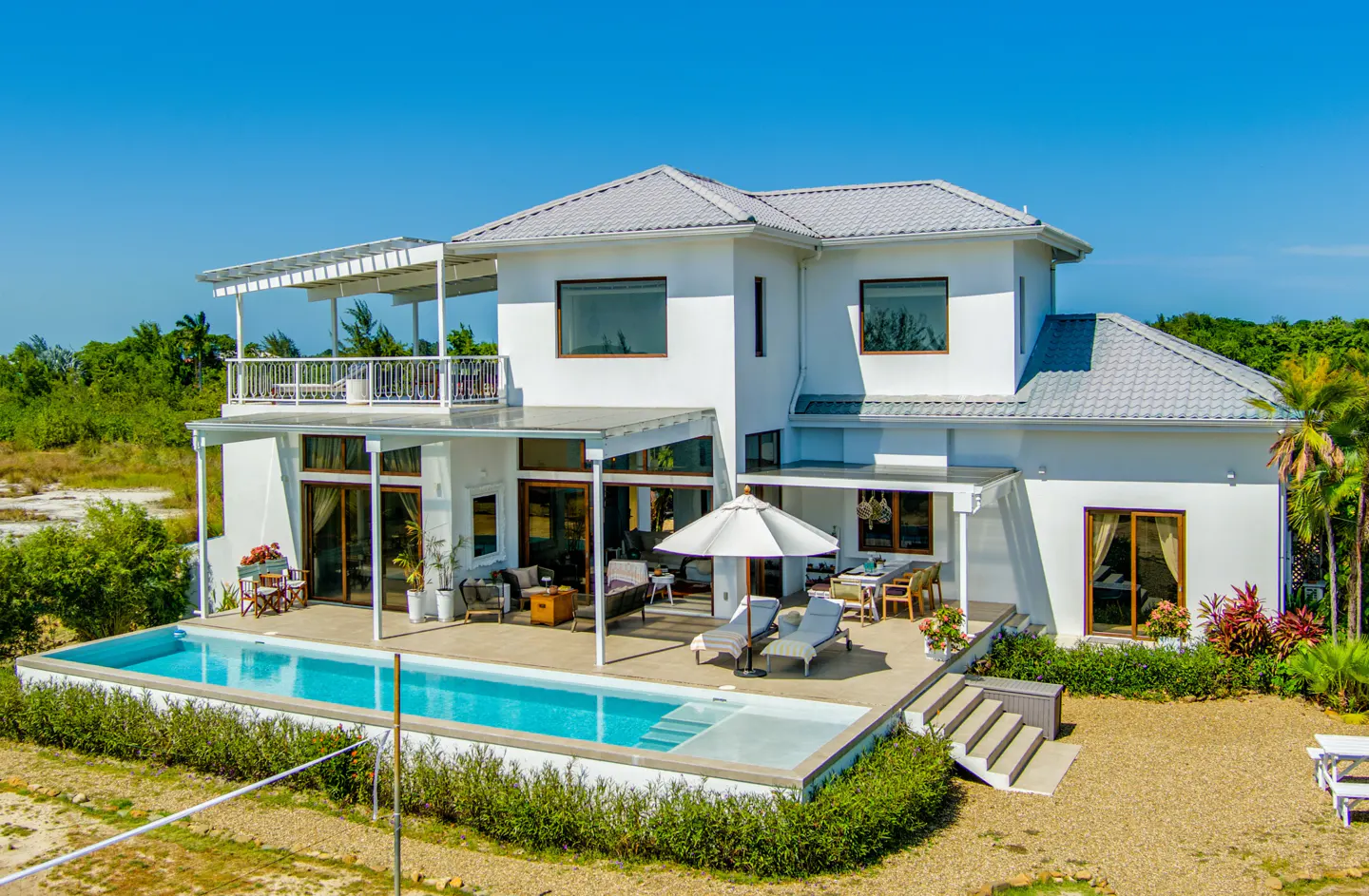 Two-story white house with a gray roof, a pool, and outdoor furniture on a sunny day.