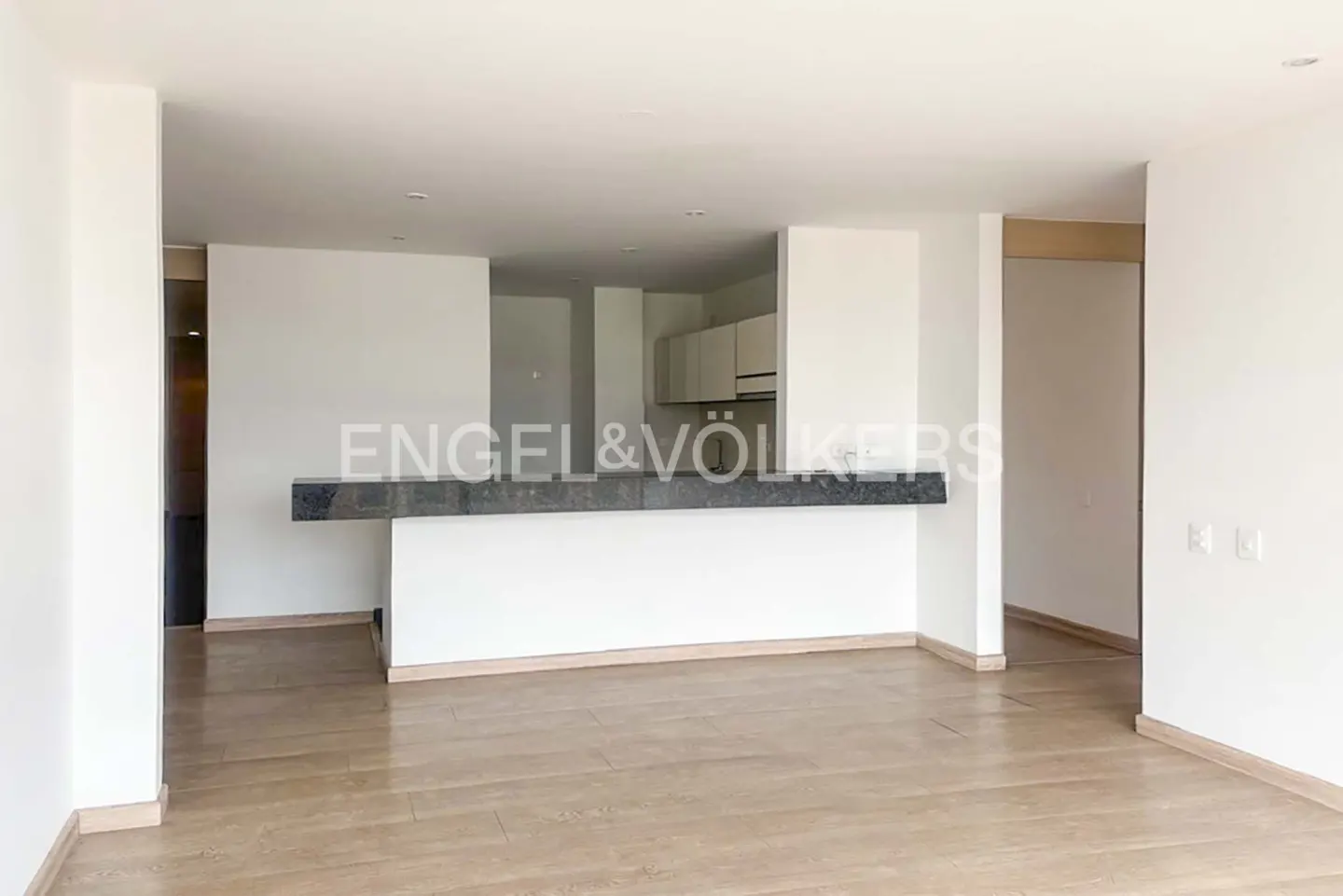 An open-concept living space with light wood floors, white walls, and a dark gray countertop bar separating the kitchen. Engel & Völkers logo is visible.