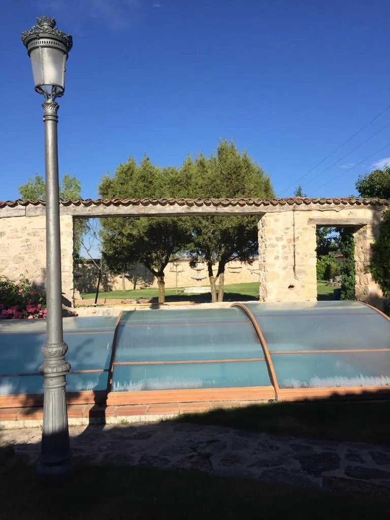A covered swimming pool sits in front of a stone wall and lush green trees under a clear blue sky, with a tall lamp post.