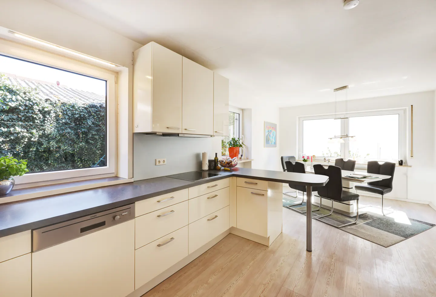 Bright kitchen with cream cabinets, dark countertops, and wood floors. A dining table with black chairs is visible in the background. A large window overlooks green foliage.