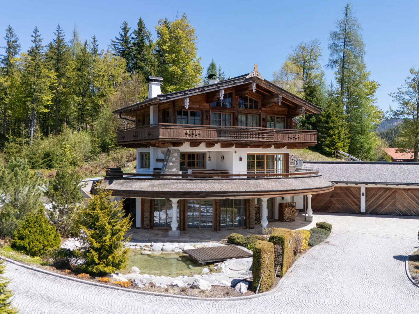 A three-story chalet-style home with a wood and white exterior, surrounded by trees and a stone driveway. A small pond is in the front.