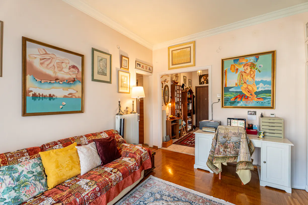 Cozy living room with eclectic decor: colorful sofa, paintings, desk with computer, and a glimpse into a book-filled room.