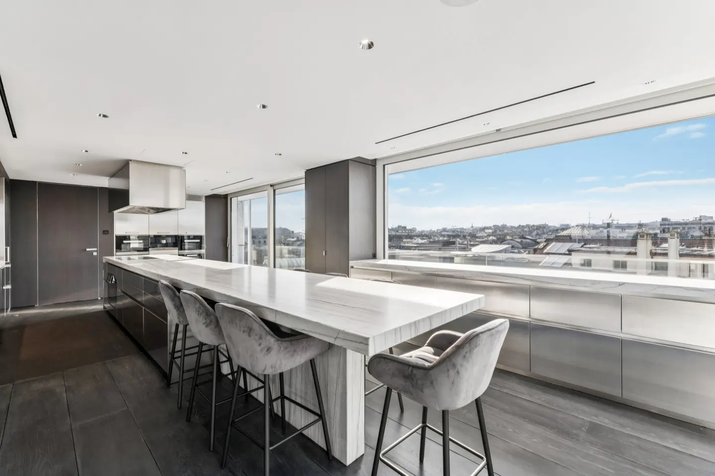 A modern kitchen with a long marble island and gray velvet chairs. A large window overlooks a city skyline.