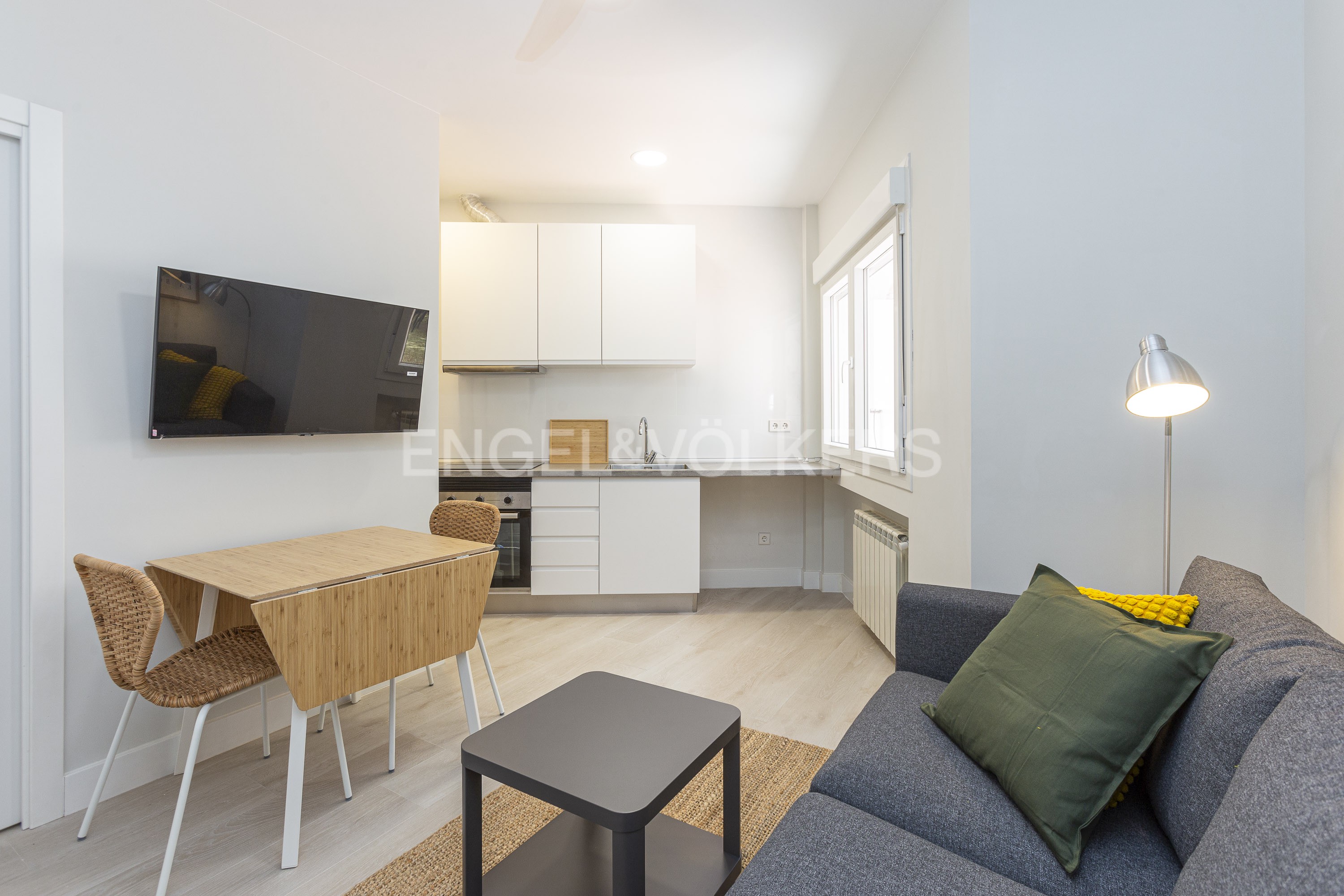 Modern studio apartment with a grey sofa, small black coffee table, bamboo dining set, and a white kitchen in the background. [AI-generated text]