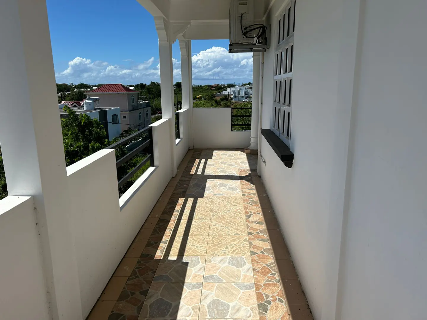 A bright, sunlit balcony with patterned tile flooring, white walls, and a black metal railing overlooking a green landscape.