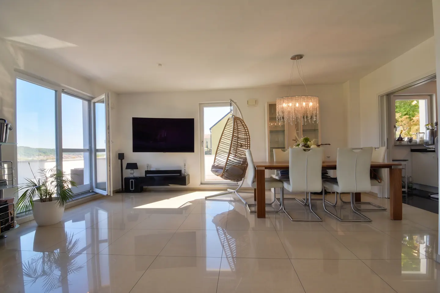 Bright, modern living room with a TV, hanging chair, dining table, and large windows. The floor is tiled and reflective.