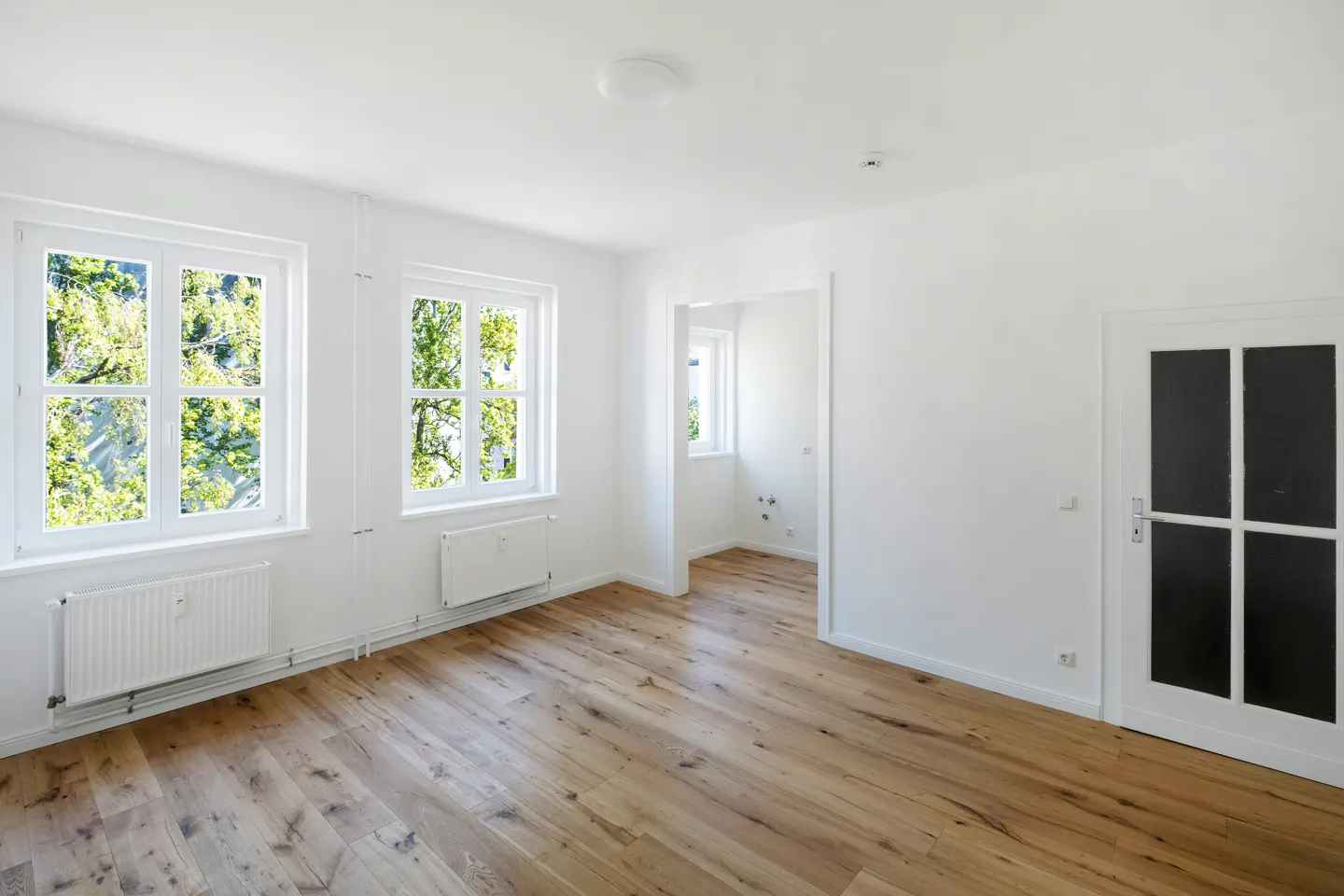 Bright, empty room with hardwood floors, white walls, two windows with green trees outside, and a white door with black panes.