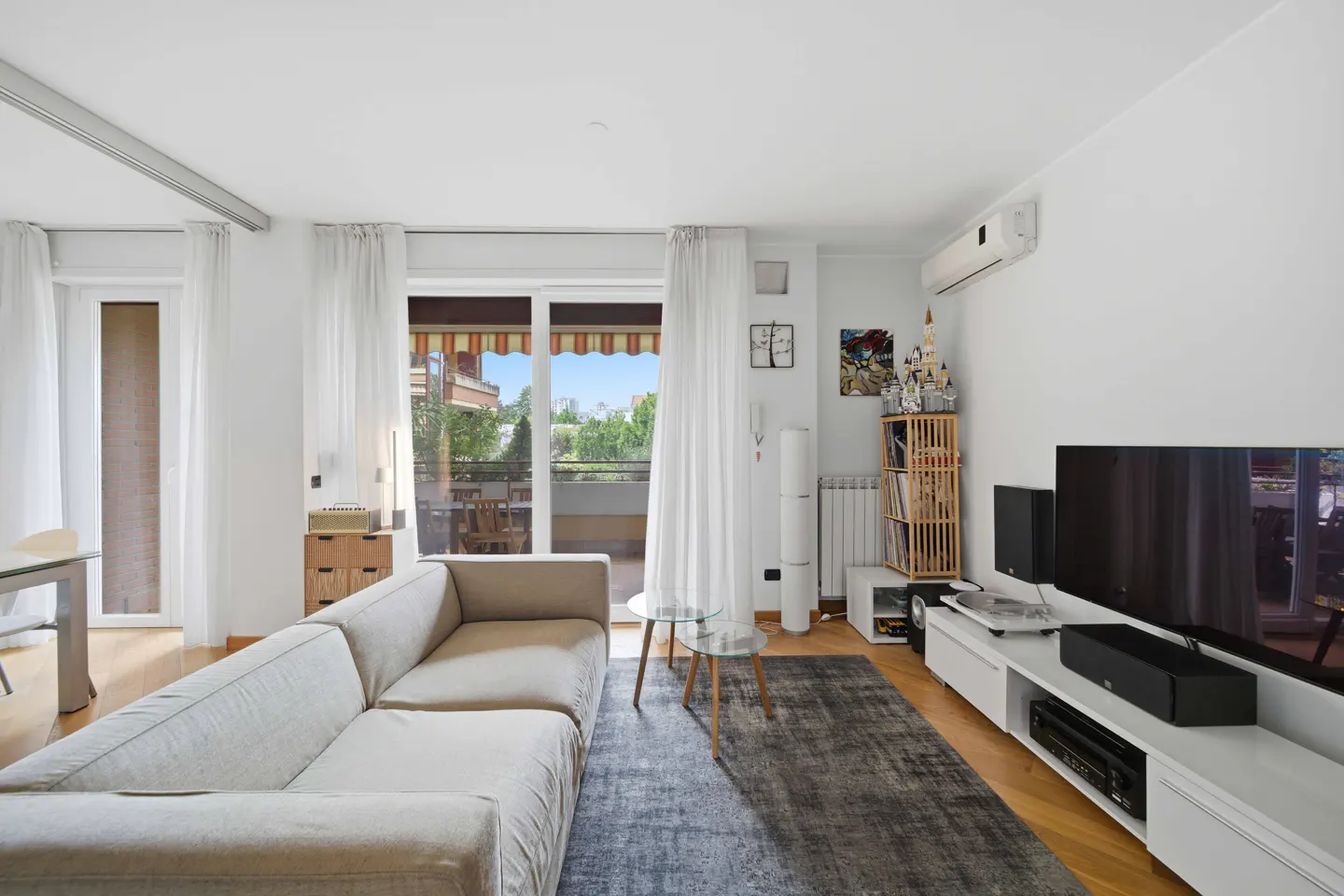 Bright living room with a beige sofa, gray rug, and white media console with a large TV. Balcony doors with sheer curtains let in natural light.