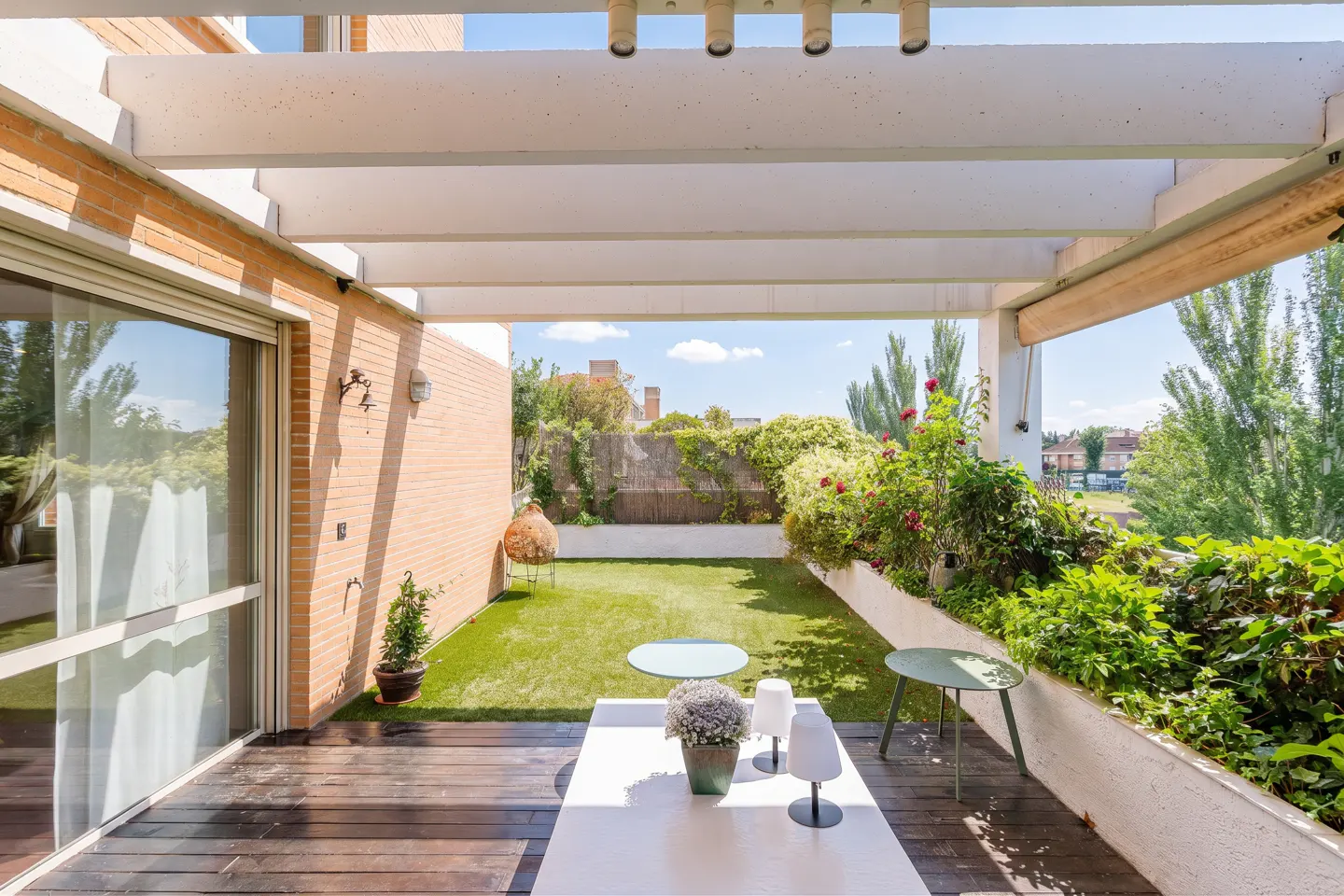 Outdoor patio with white pergola, wood deck, and green lawn. White table with lamps and flowers. Brick building and trees in the background.
