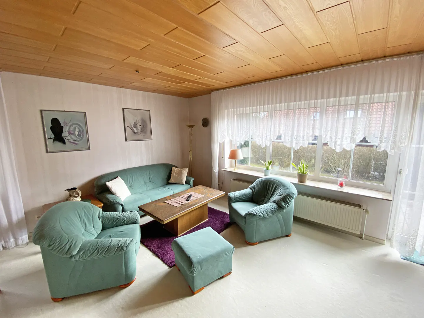 Living room with green sofa, armchair, ottoman, and wood coffee table on a purple rug. Large window with white curtains.
