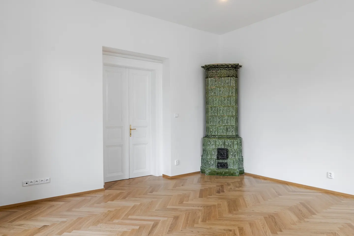 Bright, empty room with herringbone wood floors, white walls, and a green tiled stove in the corner. A white door is on the left.