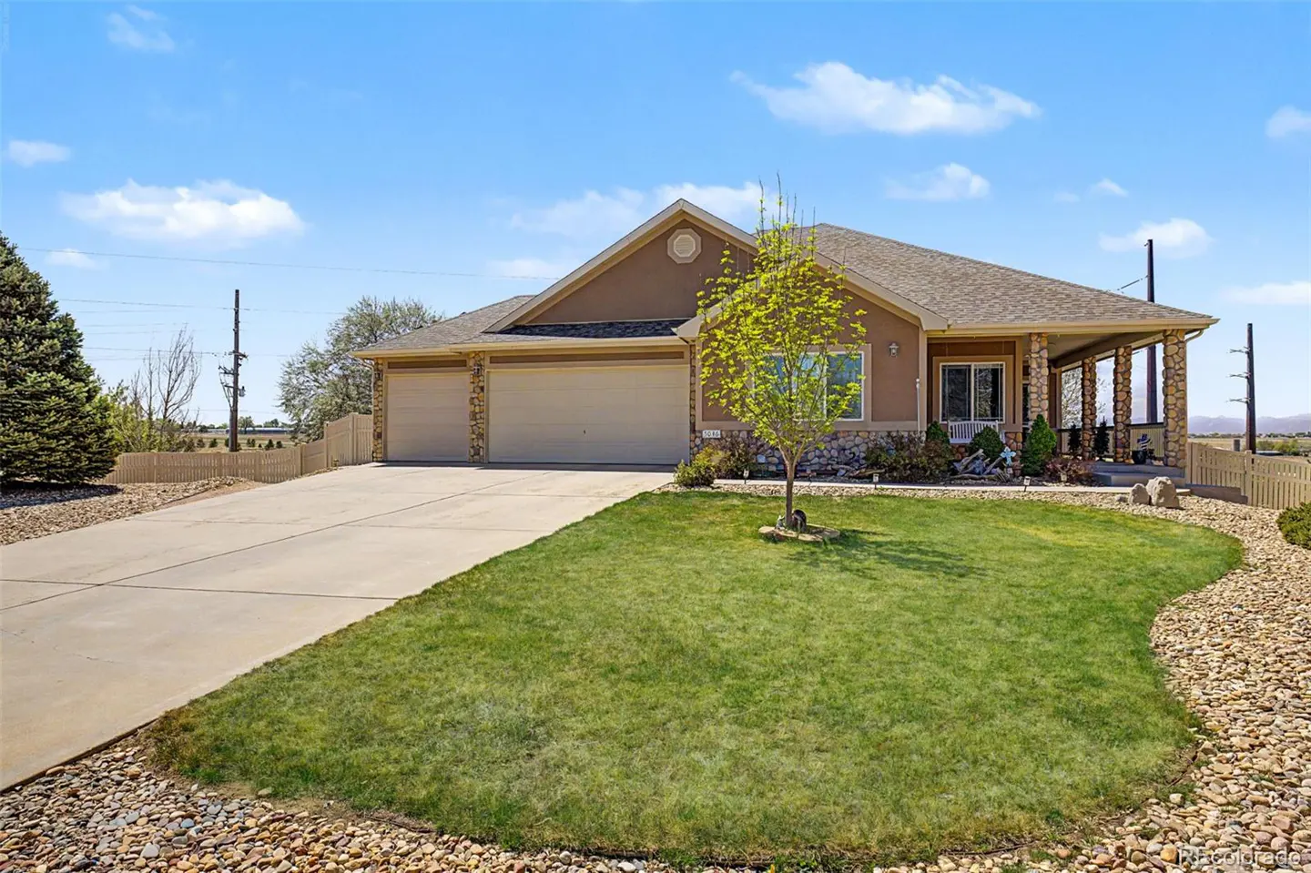 Tan single-story house with a two-car garage, green lawn, and stone accents under a blue sky.