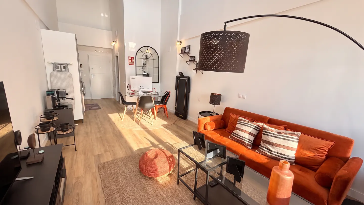 Bright, modern apartment interior with orange sofa, glass table, and dining area. Sunlight streams across the wooden floor.