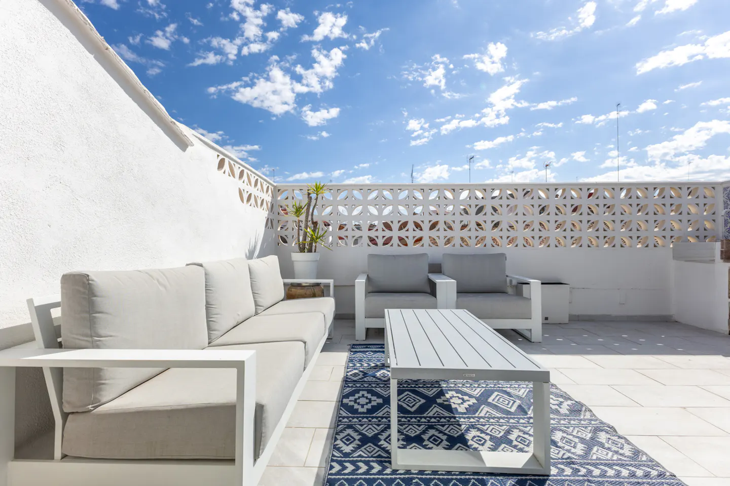 Bright rooftop patio with white furniture, a blue patterned rug, and a white decorative wall under a blue sky.