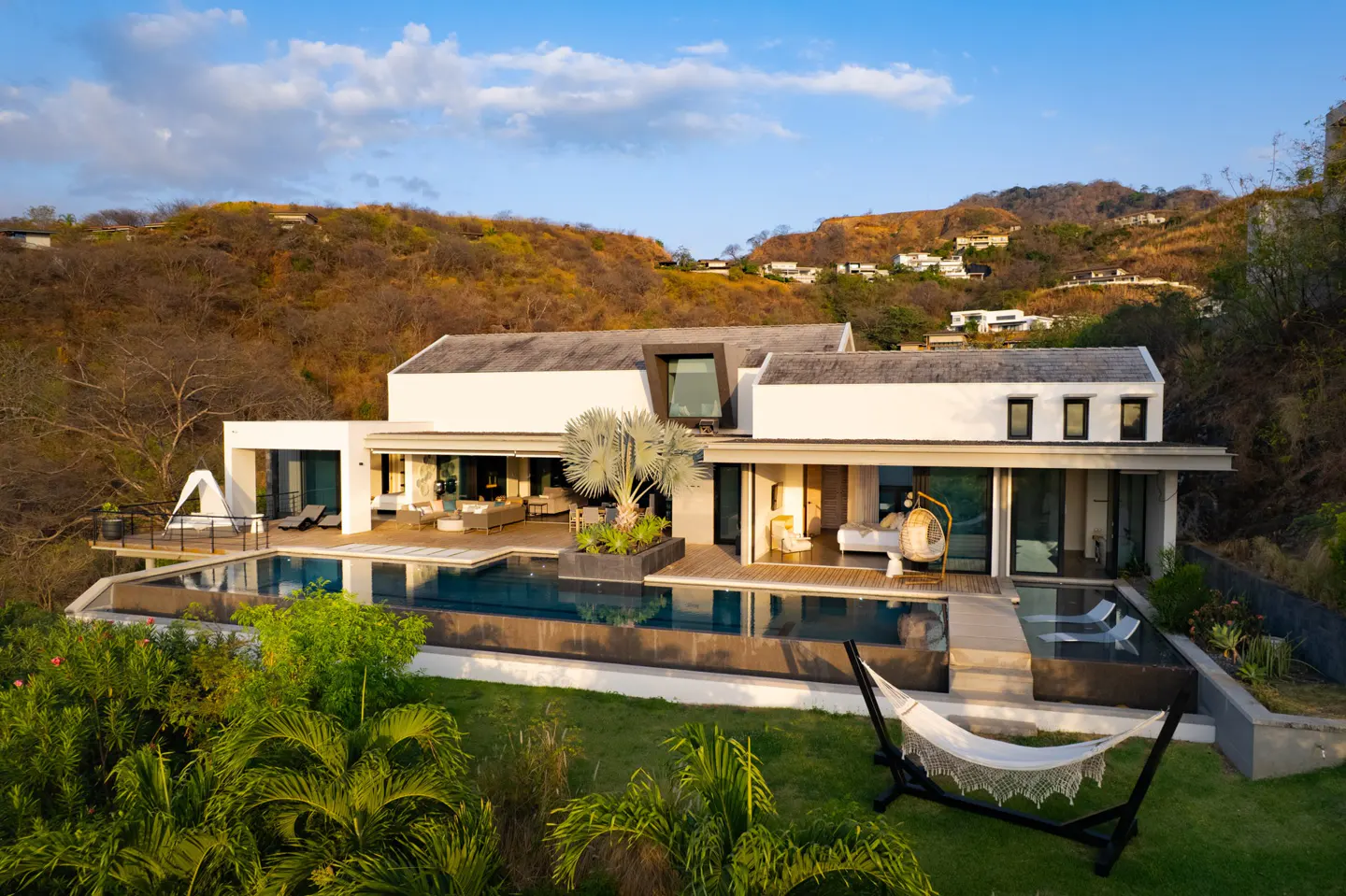 Aerial view of a modern white house with a pool, deck, and hammock, set against a hillside with scattered houses.