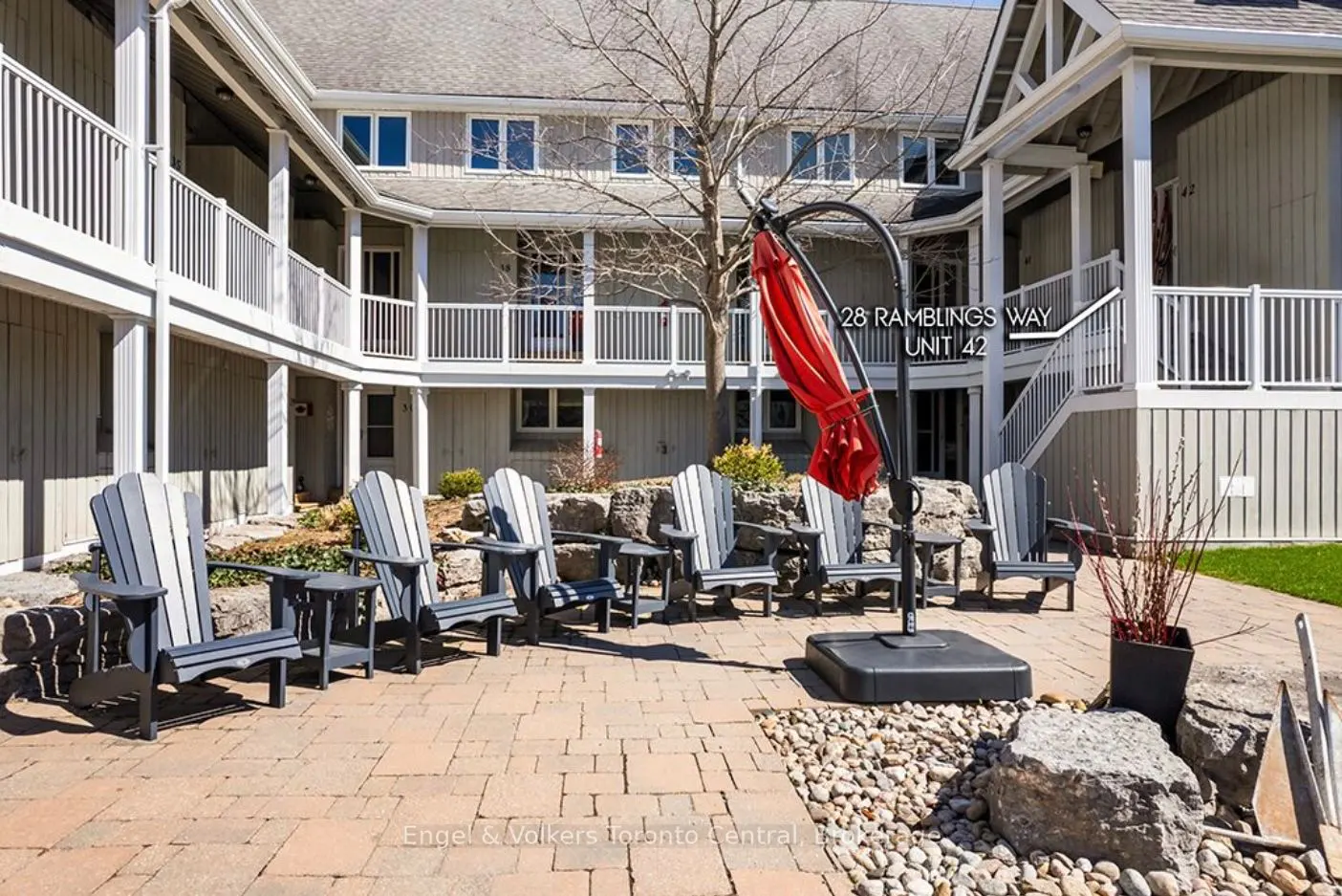 Outdoor patio with gray Adirondack chairs, red umbrella, and stone landscaping in front of a two-story building with white railings.