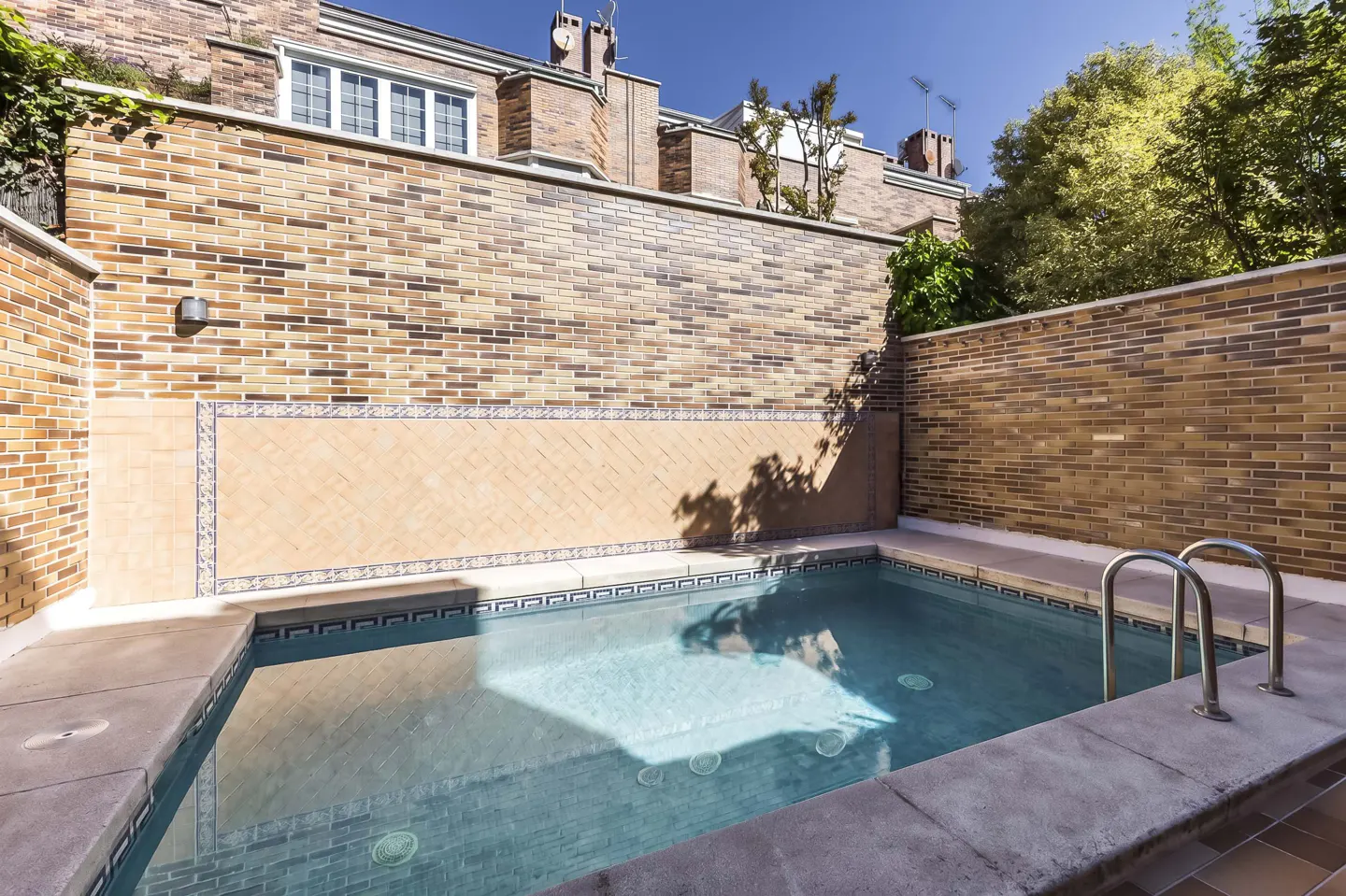 Outdoor pool with blue water, surrounded by brick walls and stone coping. Metal ladder for pool access. Building visible in background.