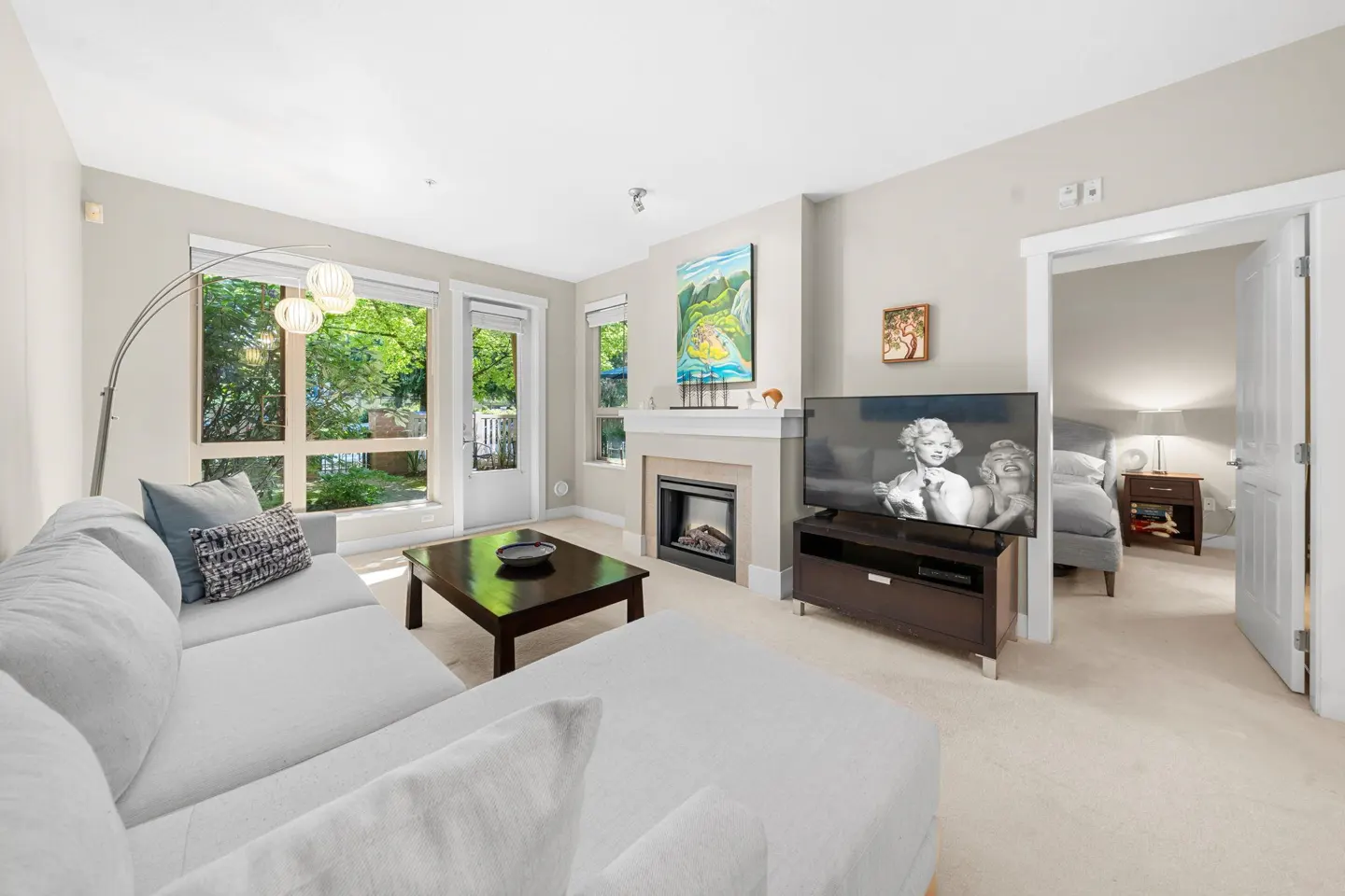 Bright living room with a light gray sectional, dark coffee table, fireplace, TV, and a view into a bedroom.