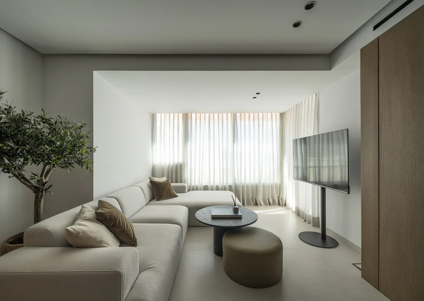 Modern living room with a beige sectional sofa, round coffee table, ottoman, and a wall-mounted TV by sheer curtains.