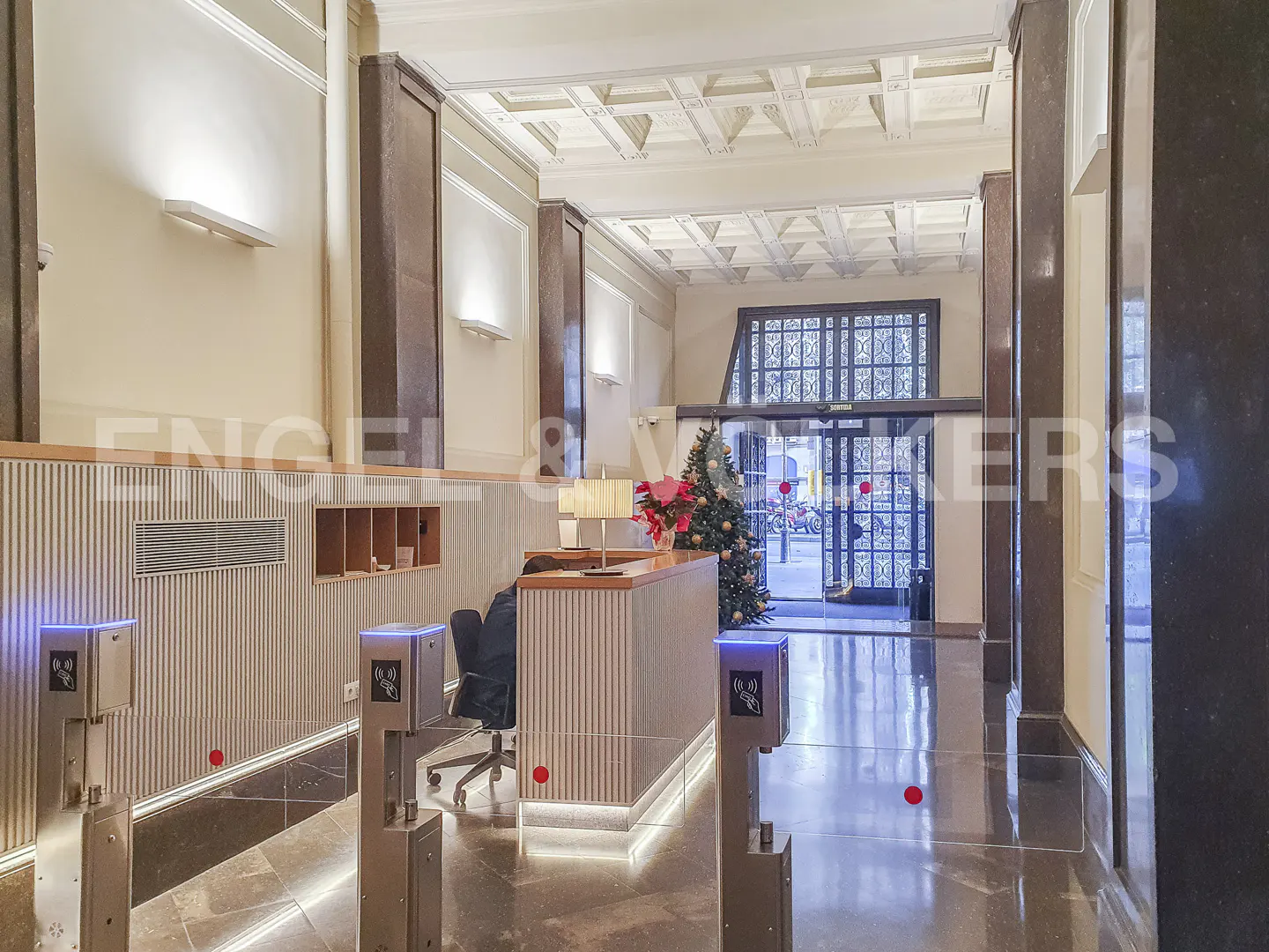 Lobby view with a reception desk, Christmas tree, and security gates. The walls are beige with brown pillars and a decorative white ceiling.